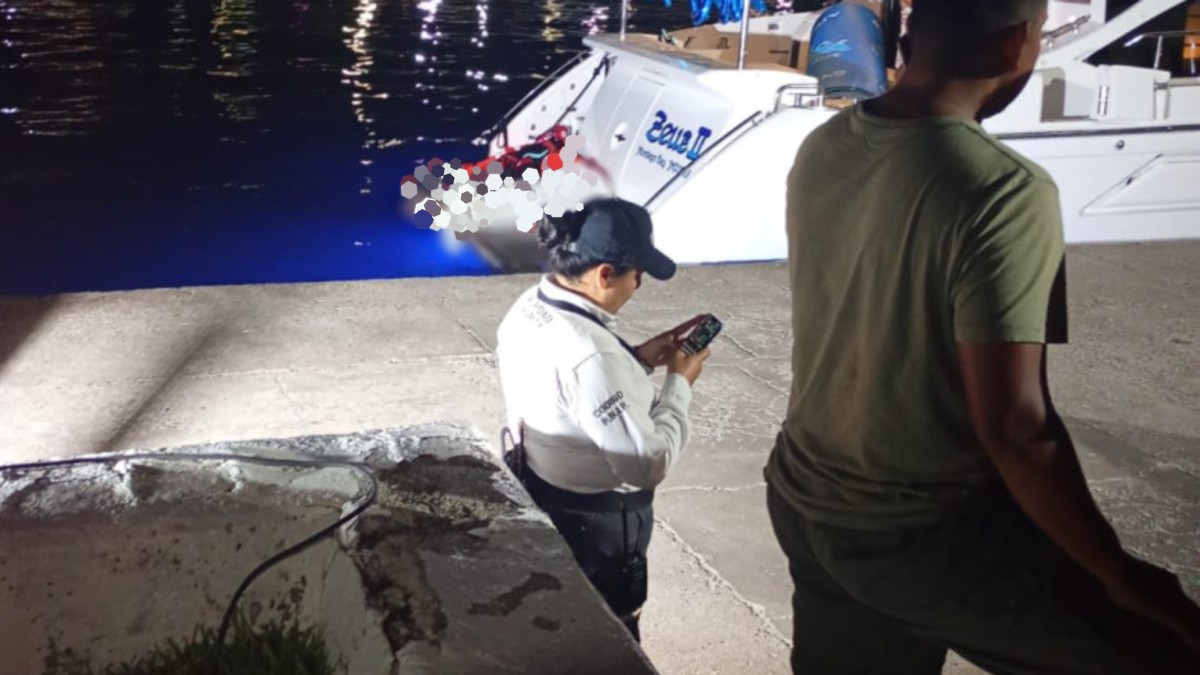 Oficial de la Policía de Puerto Vallarta sosteniendo su celular con un hombre a su derecha parados al lado de un yate a la orilla de un puerto