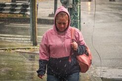 Una mujer camina bajo la lluvia en una zona urbana, vistiendo sudadera con capucha y cargando un bolso, mientras el pavimento mojado refleja las condiciones cli