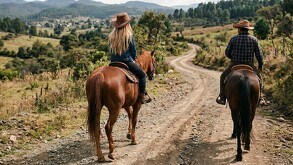 Se observa a una mujer y un hombre montando cada uno un caballo, sobre terracería.