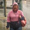 Una mujer camina bajo la lluvia en una zona urbana, vistiendo sudadera con capucha y cargando un bolso, mientras el pavimento mojado refleja las condiciones cli