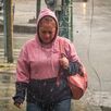 Una mujer camina bajo la lluvia en una zona urbana, vistiendo sudadera con capucha y cargando un bolso, mientras el pavimento mojado refleja las condiciones cli