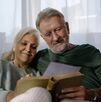 una pareja de adultos mayores, una mujer y u hombre de la tercera edad sentados en una sala mientras leen un libro