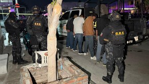 Se observa a varios policías con uniforme, hay 4 personas esposadasfrente a una patrulla; es de noche.