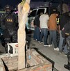 Se observa a varios policías con uniforme, hay 4 personas esposadasfrente a una patrulla; es de noche.