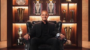 Javier Hernández, el Chicharito, vestido de negro sentado en un sillón con trofeos detrás.