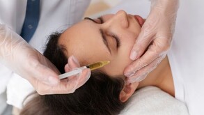 Mujer con los ojos cerrados siendo inyectada en el rostro con una jeringa con plasma sanguíneo color amarillo sostenido por unas manos con guantes