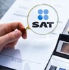 Mano sosteniendo una lupa con el logo del SAT delante de un documento con una calculadora, monedas y unos lentes a un lado