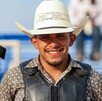Se observa a un hombre joven que porta un sombrero vaquero y está sonriendo, hay un moño negro a lado.