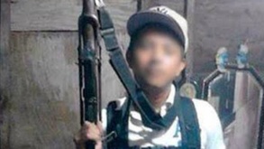 Niño con una gorra posa para una foto con un arma de grueso calibre entre sus manos.
