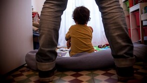 Persona parada detrás de un niño sentado en el piso sosteniendo un muñeco de bebé en un cuarto con diversos juguetes y libros alrededor