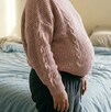 Se observa a una mujer embarazada que está en un cuarto con una cama al fondo; ella viste pants gris y sueter rosa.
