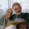 una pareja de adultos mayores, una mujer y u hombre de la tercera edad sentados en una sala mientras leen un libro