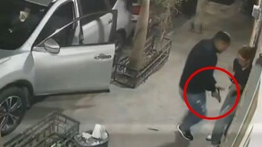 Captura de un video de seguridad con una joven siendo amenazada por un hombre con un arma de fuego al lado de una camioneta color gris con la puerta abierta