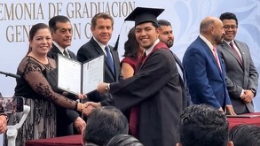 Se observa a varias personas paradas y a un joven sonriendo con una toga de graduación sosteniendo un papel en las manos.