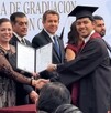 Se observa a varias personas paradas y a un joven sonriendo con una toga de graduación sosteniendo un papel en las manos.