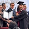 Se observa a varias personas paradas y a un joven sonriendo con una toga de graduación sosteniendo un papel en las manos.