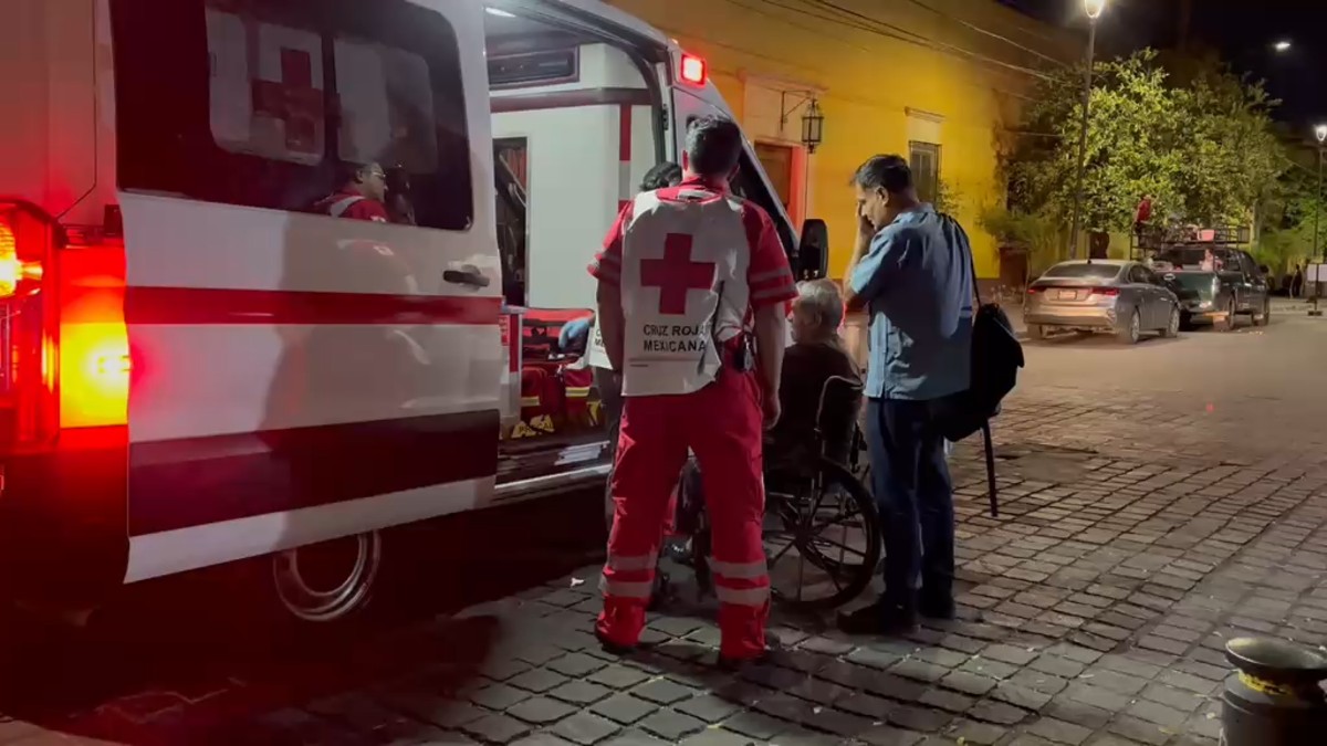 Adulto mayor en silla de ruedas con su hijo detrás, siendo atendido por paramédicos de la Cruz Roja al lado de una ambulancia en calles de Guadalajara