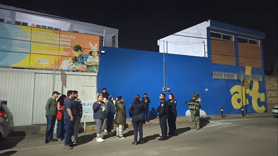 Hombres, mujeres y policías parados afuera de una escuela durante la noche.
