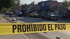 Cinta policiaca con la leyenda 'Prohibido el paso' acordonando una calle con patrullas de policía, camionetas y casas desenfocadas en el fondo