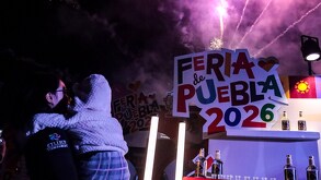 una mujer carga a un niño mientras hay fuegos artificiales y el logo de la feria de puebla 2026
