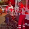 Ambulancia y tres paramédicos de la Cruz Roja atendiendo a un adulto mayor en silla de ruedas con personas y autos alrededor