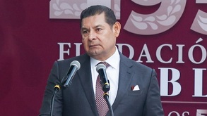 Hombre de semblante serio parado frente a varios micrófonos.