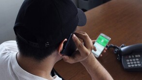 Hombre visto desde arriba vistiendo una gorra negra y sosteniendo una llamada telefónica con un celular en cada mano y un teléfono fijo al frente en una mesa