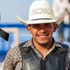 Se observa a un hombre joven que porta un sombrero vaquero y está sonriendo, hay un moño negro a lado.