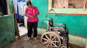 Se observa a una mujer de la terecera edad con playera rosa y agarra una silla de ruedas.