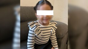 Niña con los ojos censurados por un recuadro blanco y el rostro blureado, sentada en un silló color café oscuro