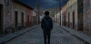 Se observa a un joven de cabello rizado con mochila, está de espaldas, va caminando en una calle solitaria.