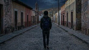 Se observa a un joven de cabello rizado con mochila, está de espaldas, va caminando en una calle solitaria.