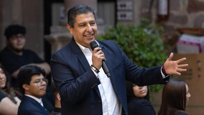 Exalcalde de Uruapan, Ignacio Campos sonriendo mientras sostiene un micrófono y durante una presentación pública con personas desenfocadas en el fondo