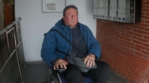 Adulto mayor identificado como Pepe sentado en su silla de ruedas con un bolso en el regazo en la entrada de un edificio