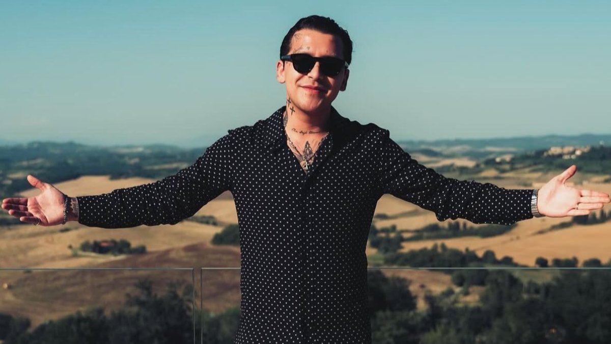 Christian Nodal está de pie con los brazos abiertos de par en par y las palmas hacia arriba, sonriendo ligeramente.