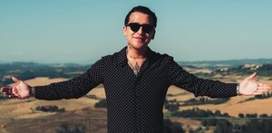 Christian Nodal está de pie con los brazos abiertos de par en par y las palmas hacia arriba, sonriendo ligeramente.