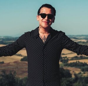 Christian Nodal está de pie con los brazos abiertos de par en par y las palmas hacia arriba, sonriendo ligeramente.