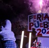 una mujer carga a un niño mientras hay fuegos artificiales y el logo de la feria de puebla 2026