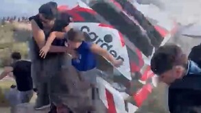 Auto deportivo participante del Rally Sudamericano en Argentina volcando al lado de una mujer y una niña con hombre huyendo alrededor