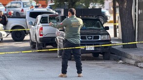 Se observa a un hombre vestido de policía, hay una cintilla amarilla de precaución y hay vehículos estacionados.
