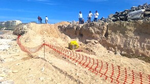 Área de construcción delimitada por una malla naranja en medio de la playa con trabajadores y personas en la parte de arriba al lado de unas rocas