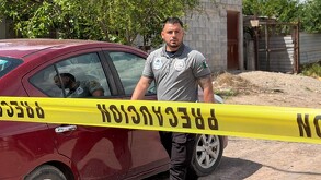 Se observa una cinta amarilla de precacuión, un hombre con brazo tatuado y uniforme de policía; atrás está un auto color rojo.