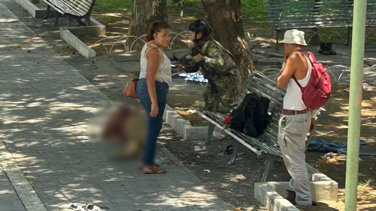 Personas paradas al lado del cuerpo de un hombre tirado sobre la acera de un parque con un militar del Ejército Mexicano cruzando la zona