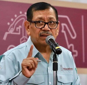 Hombre de lentes haciendo una seña con la mano mientras habla ante un micrófono.