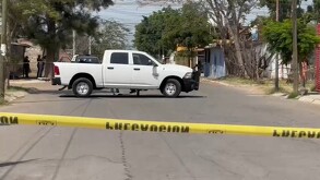 Camioneta blanca estacionada en medio de una calle acordonada por una cinta policiaca con casas, árboles y personas alrededor