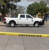 Camioneta blanca estacionada en medio de una calle acordonada por una cinta policiaca con casas, árboles y personas alrededor