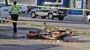 Motocicleta color naranja con negro destrozada y tendida sobre el pavimento en un área acordonada con una agente de la Policía Vial a un costado