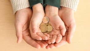 Manos de un adulto sosteniendo las manos de un niño que sostiene un puño de euros con una mesa de madera de fondo