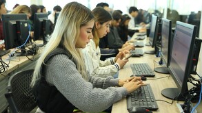 Alumna de la Universidad de Guadalajara tecleando en una computadora con grupos de estudiantes y demás computadores en el fondo