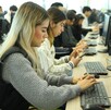 Alumna de la Universidad de Guadalajara tecleando en una computadora con grupos de estudiantes y demás computadores en el fondo